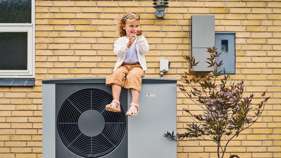 Lille pige sidder på en varmepumpe foran et gult hus